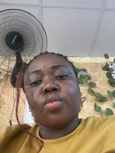 A close-up, low-angle indoor selfie depicts a dark-skinned adult female, her face prominent, looking directly at the camera. Her lips are pursed, and a small metallic stud is visible in her right nostril. Her braided hair includes reddish-brown accents, and she wears a mustard-yellow, round-neck garment.

The background shows an interior space consistent with a residence or commercial establishment within Abuja, Federal Capital Territory, Nigeria. In the upper left, a black "Century" brand stand fan is partially visible. The wall behind the subject features a light brown and tan patterned finish, possibly wallpaper or tile, with an organic, abstract design. To the right, artificial green leafy vines are affixed to the wall, alongside a decorative item composed of patterned fabric and reflective metallic strands. The upper background reveals a light grey or white ceiling or upper wall section with a visible structural seam.