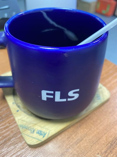 A close-up shot shows a dark purple mug with the white lettering "FLS" on its side. The mug rests on a wooden coaster with faint blue text that appears to be "Anton Krooshkin" written in a script font. Inside the mug, a white straw is visible, extending upwards and out of the frame. The mug and coaster are placed on a wooden table, with the background showing a blurred office or indoor environment. The location is not specified in the image or its context.