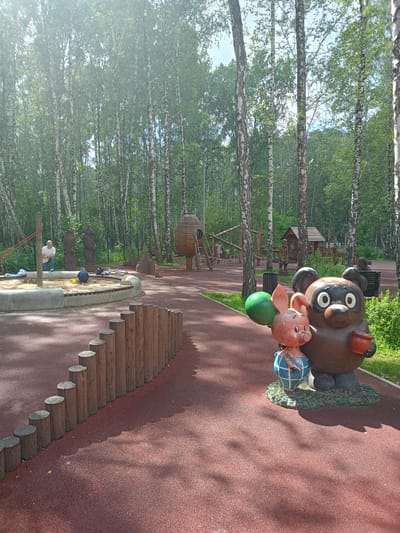 The image depicts a playground situated in a birch forest, likely in Chelyabinsk, Russia. In the foreground, a stylized statue of a pig character holding a green balloon stands next to a larger statue of a bear character holding a pot. The playground features a sandpit with a low wooden border and concrete retaining wall, where two children and an adult are visible. Further back, there is a wooden playground structure resembling a large egg with a ladder, and other wooden play elements. The ground is covered with a red, rubberized surface, and a fence made of vertical wooden logs lines the edge of the playground area. The background is dominated by a dense forest of birch trees with lush green foliage.