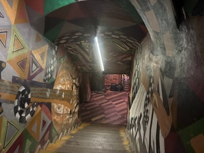 An underground passage in Sibiu, Romania, depicts a descending wooden staircase with worn, yellow-painted edges. The walls and arched ceiling are entirely covered in vibrant, abstract murals featuring geometric shapes, triangles, swirling lines, and black-and-white checkerboard patterns. A single linear fluorescent light fixture illuminates the arched ceiling, reflecting off its painted surface. At the base of the stairs, a room with a herringbone-patterned wooden floor is visible. Objects present include a wheeled cart, two black crates, and two metal kegs positioned against an exposed brick wall. The scene is devoid of individuals or activity.