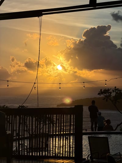 The scene depicts a sunset over a large body of water, likely a sea or ocean, viewed from an elevated vantage point in the city of Sakti, Indonesia. The sky is dominated by golden-orange hues, with the sun appearing as a bright, circular disc partially obscured by clouds in the upper-middle left, from which visible rays of light emanate. Various clouds are scattered, including a large, dark cumulus cloud silhouetted against the bright sky on the upper right.

In the foreground, structural elements are predominantly silhouetted. Dark horizontal beams span the top of the frame. A dark, beaded chain hangs vertically from an unseen point above. A string of at least five incandescent bulbs, appearing unlit, is stretched horizontally across the mid-ground, above the water. A dark, vertical-slatted wooden or bamboo railing runs across the lower-middle portion of the image, separating the viewing platform from the water. The water below reflects the golden light, creating shimmering patterns visible through the railing.

On the right side of the frame, three silhouetted human figures are observed standing, facing away from the viewer, and looking towards the sunset and the water. The leftmost figure is an adult, positioned alongside two smaller figures to their right, consistent with children or shorter adults, also observing the vista. Partial views of dark outdoor furniture, possibly lounge chairs, and a silhouetted plant with distinct leaves are visible in the lower right foreground.