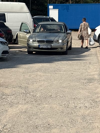 In Kobyla Góra, Poland, on a sunny day, a woman in a leopard-print dress walks away from a silver Jaguar sedan with its driver's side door open. The car is parked on a gravel lot. A white van is visible to the left, and a blue shipping container is in the background.