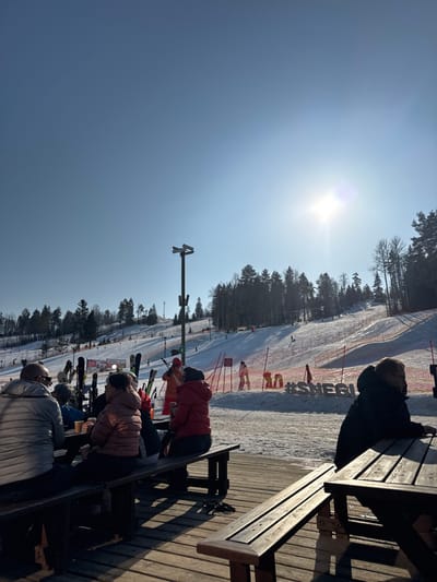 The scene depicts an outdoor dining area at a ski resort, specifically Sviyaga Hills, located in Vasil'evo, Russia, on a clear, sunny winter day. In the foreground, a wooden deck features several picnic-style tables and benches. At the left table, four individuals are seated. A man in a light gray puffer jacket and sunglasses sits with his back to the viewer. Adjacent to him, another person wears a brown/rose puffer jacket. A third, partially obscured individual is in a dark jacket, and a fourth person in a red puffer jacket holds a cup. Several pairs of skis and poles are propped near this table. To the right, another wooden table has a single man in a dark jacket and glasses seated with his back to the viewer. Long shadows are cast across the wooden deck, indicating a low sun angle.
The midground features a snow-covered area between the deck and the ski slope. A tall, dark pole with two directional speakers is centrally positioned. Orange safety netting delineates the base of the ski slope. To the right of the pole, a large, white snow installation spells out "#SNEGU." Multiple figures in ski attire are visible in this area, including one person in a bright orange jacket.
The background shows a wide, snow-covered ski slope ascending a forested hill, densely populated with evergreen trees. Numerous small figures, presumed skiers or snowboarders, are active on the slope. A ski lift tower is visible towards the upper left of the slope. The sky is bright blue and clear, with the sun prominently visible as a strong flare in the upper right quadrant.