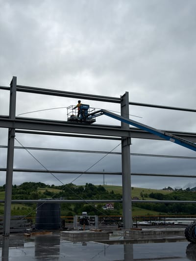 The image captures an outdoor construction site in Skrzydlna, Poland, under an overcast sky. A worker wearing a safety vest is positioned in the bucket of a blue boom lift, operating at a height among the steel framework of a building under construction. The worker appears to be manipulating a long, thin object that extends from the boom lift. The foreground shows a wet, reflective surface, possibly concrete or asphalt, indicating recent rain. In the background, a lush, green hillside dotted with houses and trees slopes upwards, with a telecommunications tower visible on the crest. Other construction materials, including a large black cylindrical tank and stacked beams, are present on the ground level.