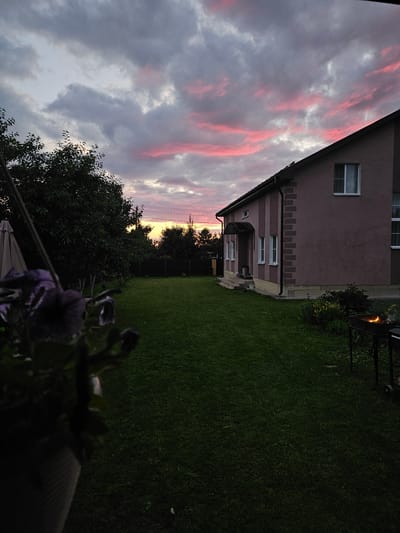 The image depicts a twilight scene in Gornye Moriny, Russia, featuring a residential house and its surrounding yard. The sky is filled with dramatic clouds, tinged with pink and purple hues, indicating sunset. A two-story house, painted a muted pinkish-brown, dominates the right side of the frame. It has multiple windows and decorative brick-like vertical accents. In the foreground, a large, dark green lawn stretches out, and on the left side, out of focus, is a potted plant with dark purple flowers. To the right of the house, a charcoal grill with glowing embers is visible. The yard is enclosed by a dark fence in the background, beyond which trees are silhouetted against the fading light. The overall impression is a serene, domestic setting at dusk.