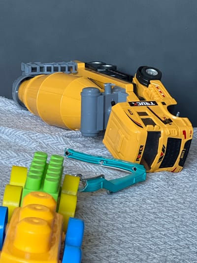 The image shows a collection of toy vehicles on a textured surface, likely a blanket or rug. A large yellow toy cement mixer truck is lying on its side. Its drum is oriented horizontally, and the truck's cab is facing towards the viewer's right. A turquoise toy excavator arm is lying detached near the cement mixer. In the foreground, several colorful toy building blocks, predominantly yellow, green, and blue, are arranged. The background is a dark, plain color, possibly a wall or backdrop. There are no people visible in the image. The scene is indoors. The image does not contain any information about the city of Aqmol, Kazakhstan.