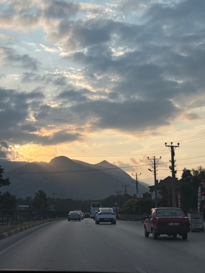 The image displays an outdoor scene in Döşemealtı, Turkey, at either sunrise or sunset, viewed from inside a vehicle traveling on a road. The sky is predominantly covered by grey and pale blue stratocumulus clouds, with the sun partially visible behind a dark mountain range on the left, casting a golden-orange glow through the clouds and across the upper atmosphere.
The foreground features a multi-lane asphalt road, with yellow painted curbs marking its left edge. Several vehicles are observed traveling away from the viewer. In the right foreground, a red, older-model sedan is visible from the rear, displaying a Turkish license plate "07 ZC 631 54". Further ahead in the left lane, a white, modern-style sedan is present, followed by a silver sedan. Additional vehicles, including a white bus, are discernible in the distance on the right side of the road.
On the right side of the road, multiple wooden utility poles equipped with cross-arms support numerous overhead power and communication lines. Behind these poles, low-rise structures with red-tiled roofs are visible. The background is dominated by a range of dark, silhouetted mountains. Sparse vegetation, consisting of trees and bushes, lines the road and is scattered throughout the landscape. No human figures are clearly identifiable.