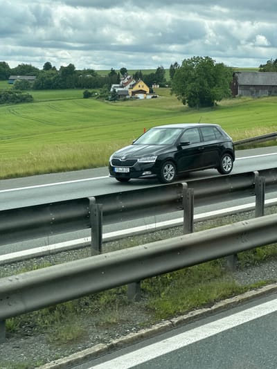 A black Skoda Fabia hatchback, license plate FB-JO 32, is driving on a multi-lane highway under a cloudy sky. The car is in the right lane. The highway is bordered by metal guardrails. In the background, there is a rolling green landscape with fields, trees, and several houses, including a yellow house with a red roof and a large barn. The scene appears to be in a rural area outside the city of Köditz, Germany, based on the license plate format and the landscape. The weather is overcast.