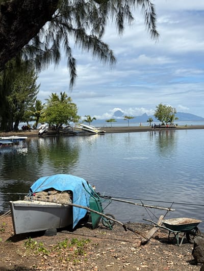 The image depicts a tropical coastal environment in Māhina, France, under a partly cloudy sky. In the foreground, a small, grey, flat-bottomed boat, partially covered by a blue tarp revealing mesh bags beneath, rests on dark, reddish-brown ground with sparse vegetation. To its right, an olive-green two-wheeled cart and a rusty single-wheeled wheelbarrow are situated near the calm, dark water. Several long poles, consistent with fishing rods, extend from the foreground towards the water. The opposite shore, composed of dark sand, hosts tropical vegetation including palm and broadleaf trees. Multiple white and yellow kayaks or outrigger canoes are pulled onto this shore, where a small group of indistinct individuals is visible standing. Distant, cloud-shrouded mountains form the background.