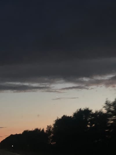 The image shows a sky with clouds during dusk or dawn, with a silhouette of trees in the foreground. There are two small points of light visible in the sky, one brighter than the other. The sky transitions from dark gray clouds at the top to lighter hues of orange and pink near the horizon. The trees are dark and appear to be in motion, possibly due to the camera's movement. There is no clear indication of the city of Акылбай, Kazakhstan, or any specific landmarks within the image.