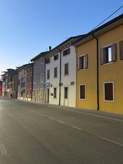 The image shows a street in Villafranca di Verona, Italy, lined with buildings of varying colors and heights. The buildings are adjacent to the road. The sky is visible on the left side of the image. A street stretches towards the horizon, indicating the linear layout of the town. A person can be seen walking along the street further away in the distance.