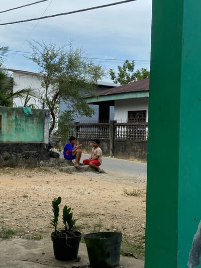 The image displays an outdoor scene, likely in Gampongaceh, Indonesia, framed by a green architectural element, possibly an overhang or balcony, at the top and bottom. Two young boys are central to the image, crouching on a concrete ledge next to a dirt and gravel path. The boy on the left wears a blue shirt and shorts, looking towards the other boy. The second boy, on the right, is in a light-colored shirt and red shorts, also crouching and facing the first boy, suggesting a shared activity or conversation.

In the background, there are houses and buildings, including a white house with a dark, patterned fence and another structure with a weathered greenish-blue wall. A paved road or path is visible behind these buildings. A green leafy tree stands on the left, reaching towards a bright, slightly cloudy sky, with several utility lines visible against it. In the foreground on the right, there are two black potted plants, one containing a leafy green plant. The scene depicts a casual, ordinary moment during what appears to be a clear, sunny day. No visible text is present.