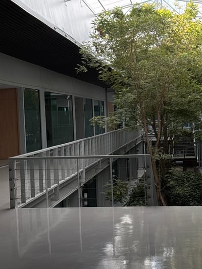 A modern building interior view includes a long, silver-colored metal staircase extending upwards. A tree with light green foliage stretches out above the staircase. Sections of the building feature large, rectangular windows and dark paneling. Lush green plants hang from the building's architecture. A textured, gray surface occupies the right side of the image, possibly a wall or reflective panel. The perspective is tilted, looking upwards at an angle.