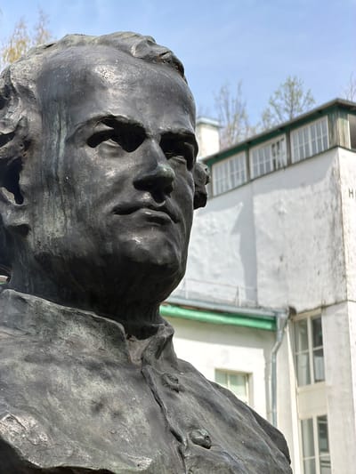 A low-angle, close-up shot of a dark metal sculpture of a human head in an outdoor setting. The head, with deeply recessed eyes and distorted features, appears to be tilted back. The rough, textured surface of the sculpture contrasts with the smooth, overcast sky visible in the background. Behind the head, slightly out of focus, is a two-story white building with multiple rectangular windows and a green stripe. The location is the city of Pavlovo, Russia.