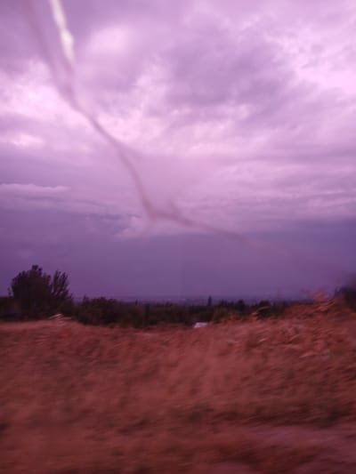 The image captures a hazy, purple-hued sky with clouds. In the foreground, a field of dry, reddish-brown grass is visible, appearing blurred due to motion. A line of dark trees forms a distant horizon, behind which a faint, indistinct city skyline can be seen. The overall impression is of a natural landscape under a dramatic sky, possibly during twilight or a storm. No people or specific landmarks are identifiable. The image is taken from a moving vehicle, as indicated by the motion blur. The location is specified as Auk, Kyrgyzstan.