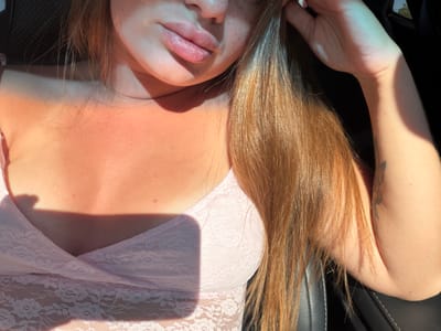 The image shows a close-up, waist-up view of a person with fair skin, freckles, and long, auburn hair. They are wearing a light pink lace top. The person is inside a vehicle, with the interior visible as a dark seat and part of the window or a black background. Sunlight casts strong shadows across the person's chest and shoulder. There is a faint tattoo on the person's arm. The environment suggests an outdoor setting, possibly during the day, given the strong sunlight and shadows. The location in Novooleksandrivka, Ukraine is not discernible from the visual information presented in the image.
