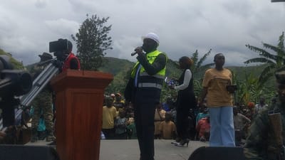 A man wearing a white hardhat, a black suit, and a yellow safety vest, stands behind a wooden podium, holding a microphone and speaking to a crowd of people in Sake, Democratic Republic of Congo. The event takes place outdoors, under an overcast sky. There is a large crowd of people, mostly adults, who are listening attentively. A cameraman stands in front of the podium, holding a large camera, and a second cameraman, holding a smaller video camera, stands behind the podium. There are several palm trees in the background. A woman stands on the left side of the speaker holding papers in her hands. She is wearing a black dress and is looking at the speaker. Another woman stands on the speaker's right, wearing a yellow shirt and blue pants. She is looking straight ahead, and holding a phone in her hand. The speaker's left hand is holding a microphone, his right hand is gesturing towards the crowd. There are many other people standing behind the crowd, some wearing camouflage uniforms. The event is taking place on a stage, which is constructed from wooden boards and has been erected on a dirt path. A video camera, with a long microphone boom attached, is in the foreground. The scene is typical of a public event, with a speaker addressing a crowd of people.