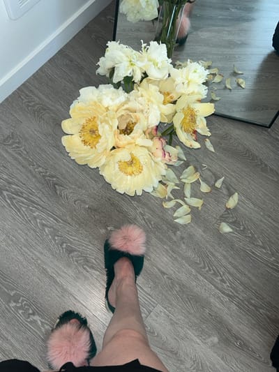 The image shows a close-up, low-angle view of a person's legs and feet, wearing dark green furry slippers with large pink pom-poms, standing on a distressed grey wooden floor. To the left, a cluster of pale yellow peonies with pink tinges are arranged, some with fallen petals scattered on the floor. A clear glass vase holding more peonies is partially visible in the upper right portion of the image. A portion of a full-length mirror is also visible in the upper right, reflecting more of the peonies and possibly another person's leg. The background shows a white wall with a thin white baseboard. The lighting suggests an indoor environment. There are no specific landmarks to indicate the city of Lynnwood, United States of America.