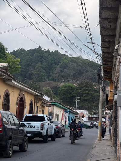 A street in La Esperanza, Honduras is captured in this ground-level photograph. Two motorcyclists, one wearing a blue shirt and the other wearing a black shirt, are riding past parked cars on a street lined with buildings. A white Isuzu pickup truck and a dark SUV with spare tires piled on top are prominently parked on the right side of the road. In the distance, a person stands on the sidewalk, holding a phone up to their ear. The backdrop is dominated by a tree-covered mountain, with numerous power lines visible against the overcast sky. Architectural details of the buildings, including arched windows and stone facades, are observable along the sides of the road.