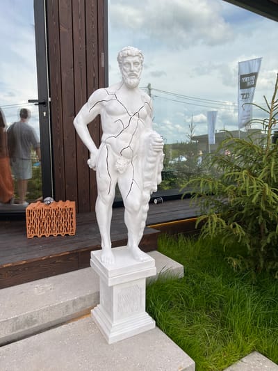 In Yakunino, Russia, a white plaster statue of a male figure, appearing to be a classical sculpture with cracked details, stands on a white pedestal in front of a modern building with dark wooden siding and large glass windows. A person's reflection is visible in the glass. To the left of the statue, on a wooden deck, is a single brick with a device on top. To the right of the statue, a small evergreen tree grows in lush green grass. The sky is overcast with clouds.