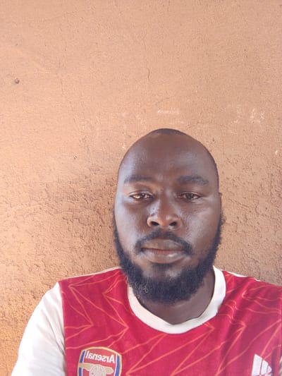 A close-up photograph shows a dark-skinned man with a shaved head and a full black beard. He is wearing a red and white soccer jersey. The background is a textured wall painted a light brown color. The location is Mulke Barikin Sayejo, Nigeria.