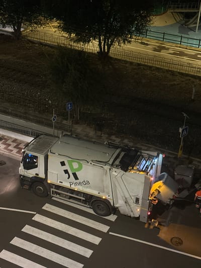 This nighttime image captures a white refuse collection truck operating on a paved street in Pineda de Mar, Spain. The truck, labeled with a green "Pineda a punt" logo, is actively emptying a large, dark grey refuse container into its rear compaction unit; the container is suspended and inverted. A sanitation worker, dressed in an orange and grey high-visibility uniform, stands to the right of the truck near an identical second refuse container.

The street features a pedestrian crossing with white zebra stripes in the foreground. Adjacent to the street, a patterned sidewalk borders an elevated embankment containing railway tracks, separated by a metal fence. Trees and sparse vegetation grow on this embankment. In the upper right background, an illuminated light blue paved area, indicative of a recreational facility, is visible. Street signs, including a pedestrian crossing symbol and a 'no entry' sign, are present along the road, which is illuminated by artificial light sources. A manhole cover is visible on the road surface near the pedestrian crossing.