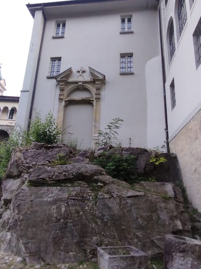 The image depicts the exterior of a building, likely a religious or historical structure, with a prominent stone facade. The building is a light, neutral color and features several windows, some with bars. Above a recessed arched entryway, a small cross is visible. In the foreground, a natural rock formation with abundant green foliage covers a significant portion of the image. Two stone planters are visible at the bottom right of the frame. The setting appears to be a hillside, with a portion of another building with arched windows visible to the left. The overall impression is of a serene, somewhat overgrown historical site. This scene is located at the Sanctuary of the Madonna del Sasso in Orselina, Switzerland.