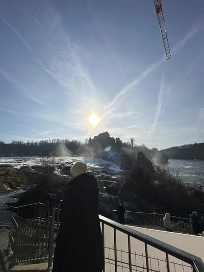 The image displays an outdoor scene at the Rheinfall in Neuhausen am Rheinfall, Switzerland, under a clear, bright blue sky. The sun, positioned in the upper-left quadrant, casts significant glare across the scene. Multiple white aircraft contrails are prominently visible, crisscrossing the sky. A segment of an orange crane boom extends downwards into the upper right portion of the frame.
The central feature is the wide, turbulent waterfall, generating visible mist, particularly on the right side. On a hill directly behind the falls, Schloss Laufen (Laufen Castle) is silhouetted against the bright sky. A dark, multi-arch bridge spans the Rhine river in the distance, positioned upstream on the far left. The riverbanks and distant hills are covered with bare trees and rocky terrain.
In the immediate foreground, a person stands with their back to the viewer, clad in a dark, long coat and a light-colored knitted beanie, observing the waterfall from a viewing platform. This platform is edged by a dark metal railing with vertical bars, partially visible on the left and lower right. Several other individuals are present further along the platform to the right, similarly observing the falls.