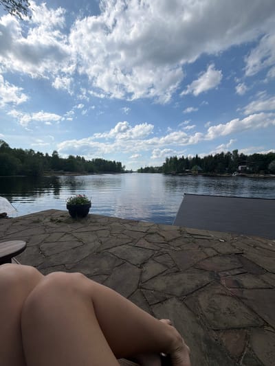 The image captures a scene from what appears to be a waterfront setting in Shchelkovo, Russia. A body of water, possibly a river or lake, stretches into the distance, bordered by dense green trees on the far bank. The sky is filled with fluffy white clouds against a blue backdrop. A portion of a deck or dock is visible, constructed from stone or paving slabs. A planter with flowers sits near the edge of the water. In the foreground, a person's legs are visible, crossed, suggesting relaxation or leisure. The overall impression is that of a calm, sunny day spent by the water.
