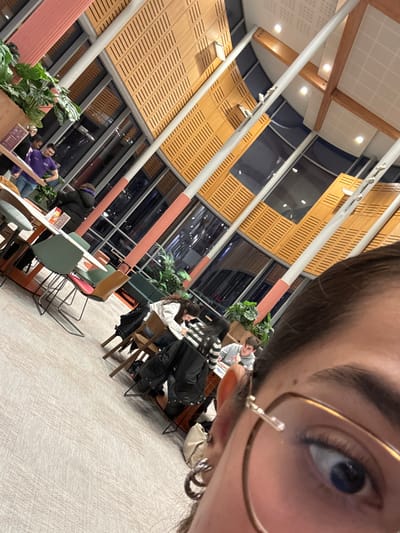 The image is a close-up selfie taken at a tilted angle, focusing on the photographer's right eye and the top portion of their face, while simultaneously capturing a wide view of a modern indoor space in the background.

In the foreground, taking up the top right quadrant of the frame, is the photographer's right eye, eyebrow, and a portion of their forehead and dark hair. The eye is a striking light blue, and the eyebrow is dark. The photographer is wearing thin-framed glasses, which appear to be a metallic gold or bronze color. A silver-toned earring, seemingly composed of multiple small rings or coils, is visible on the earlobe.

The background reveals a spacious, well-lit interior, characteristic of a contemporary public or semi-public area such as a university common room, a large café, or a modern co-working space. High ceilings and expansive windows dominate the architectural design. Through these large windows, the outside environment is dark, indicating that the photograph was taken at night or during the late evening.

The interior design features prominent light-colored wooden slatted panels on the walls and possibly parts of the ceiling. Some of these panels have small perforations, suggesting acoustic treatment. White or light grey support columns and beams traverse the space, with some sections accented in a subtle peach or light pink hue. Recessed lighting fixtures provide warm illumination throughout the area.

Several individuals are present in the background, seated at various tables. In the mid-ground, slightly to the left of the center, two women are seated at a table. One is wearing a white long-sleeved top with a dark pattern, and the other is dressed in a black and white striped top. A dark jacket is draped over a chair beside them. Behind them, another person in a grey top is partially visible, looking towards the right. Further into the background, another individual wearing a purple top is seated at a table. The people in the background appear to be casually engaged, possibly studying, working, or socializing.

The furniture includes a mix of modern tables, some with light wood surfaces and others featuring green or pink tops. The chairs also vary in style, with some being wooden and others having metal frames with colored seats. Lush green potted plants are strategically placed throughout the space, serving as decorative elements and subtle dividers. A light-colored bag is visible near one of the tables in the mid-ground.

The overall atmosphere is relaxed and informal. No discernible text or signage is visible within the image.