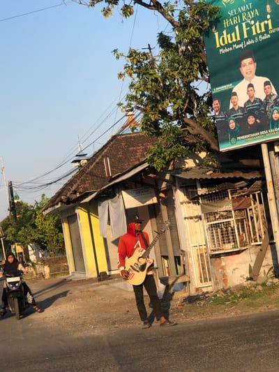 A man in a red hoodie and black pants is playing a guitar on a street in Kalidawir, Indonesia. He is holding a light-colored acoustic guitar and appears to be singing or performing. To the left of the man, a woman wearing a hijab is riding a motorcycle. In the background, there are several small shops with traditional Indonesian architecture, including tiled roofs and wooden structures. A large banner displaying a greeting for Eid al-Fitr, featuring portraits of local politicians and the text "SELAMAT HARI RAYA IDUL FITRI," is visible on the right side of the image. The sky is clear and blue, suggesting it is daytime, likely late afternoon given the long shadows. Power lines are strung across the scene, typical of many Indonesian towns. The environment appears somewhat cluttered, with debris visible along the side of the road.