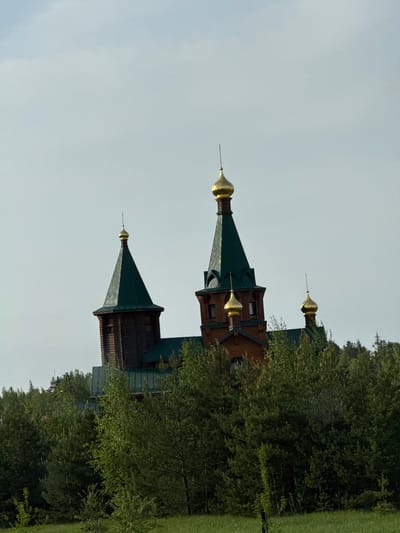 The image shows a wooden church with golden domes in Kstovo, Russia. The building has a dark brown facade and green roofs. The church is partially obscured by trees. The sky is overcast.