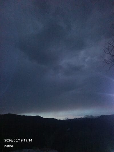The image depicts a dark, cloudy sky over a silhouetted mountain range. The time stamp "2026/06/19 19:14" and the word "natha" are overlaid on the bottom left of the image. The scene is likely a landscape shot taken during dusk or before a storm. There are no people or specific objects visible in the foreground, and the environment is characterized by darkness and the dramatic sky. The location in El Chaparral, Venezuela, is a detail provided externally and not discernable from the image itself.