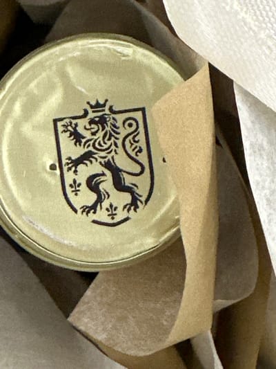 This is a close-up, high-angle, eye-level shot of a golden-colored lid with a dark emblem. The emblem is a stylized heraldic lion rampant within a shield, adorned with decorative flourishes. The lid appears to be part of a container, possibly a jar or tin, and it is partially obscured by a piece of tan paper wrapping. The background consists of soft, draped material in shades of beige and off-white, suggesting a package or wrapping. The lighting is soft and diffused, creating subtle highlights and shadows on the lid's surface, giving it a slightly textured appearance. There is a small, dark dot visible in the center of the lid, which might be a small imperfection or a deliberate marking. The scene is still and the focus is sharp on the lid and emblem. No people or discernible setting details beyond the packaging are visible. The image appears to have been taken indoors with natural or soft artificial light. There is no visible text in the image.