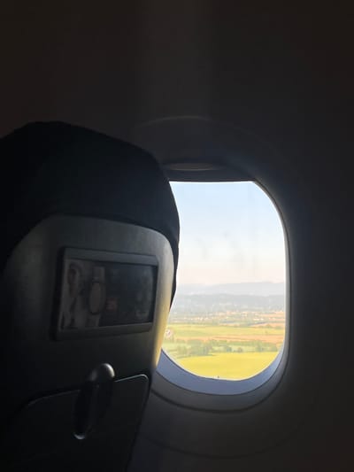 The image depicts a view from an airplane window. Outside, the landscape shows a mixture of green fields, vegetation, and possibly some urban or residential areas in the distance. The sky appears bright and clear. Inside the aircraft, the back of a seat with a screen is visible. Based on the landscape and sky, it is possible that the location could be near Colombier-Saugnieu, France, though this is not definitively proven by the image alone.