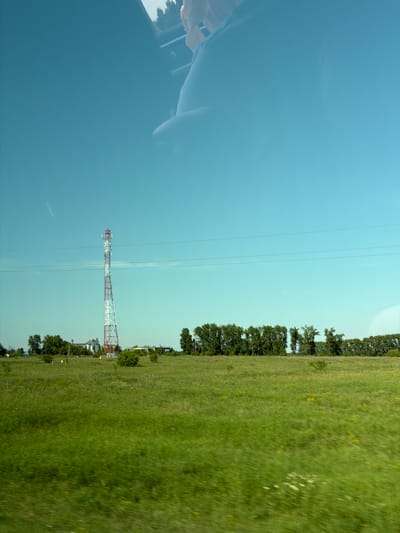 A static, eye-level medium shot captures a wide expanse of a bright, clear sky above a green, slightly blurry field. In the mid-ground, a tall, red and white lattice telecommunications tower stands prominently on the left side of the frame, its peak reaching almost to the horizon. Behind the tower, a line of trees stretches across the landscape, with several buildings visible among them. The foreground is a vibrant green field, with some out-of-focus vegetation scattered throughout. The image appears to have been taken from a moving vehicle, as indicated by the slight motion blur in the field and the reflection of a person's arm and head in the upper portion of the frame, suggesting it was taken through a car window. The overall scene depicts a rural or semi-rural setting.