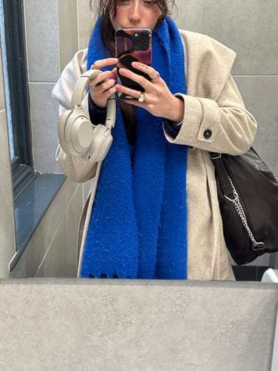 A mirror selfie reveals a person wearing a beige coat and a large, fluffy blue scarf. A black handbag with a silver chain strap rests on the coat. The person holds a phone in one hand and a pair of beige headphones in the other. Multiple rings adorn their fingers. The selfie captures the person's face partially, showing dark hair and some facial features. The background includes grey tiling.