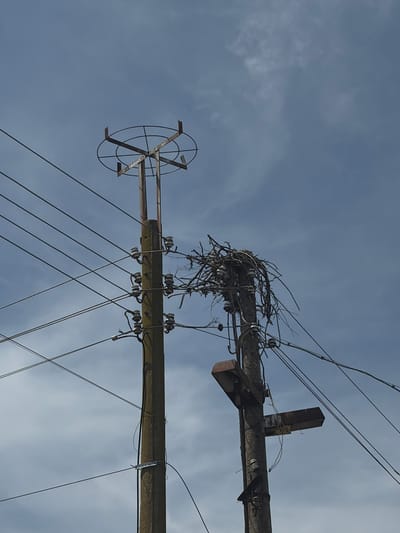 The image shows a weathered utility pole against a cloudy sky. The pole has multiple horizontal arms with ceramic insulators supporting electrical wires. A large bird's nest is situated on one of the upper arms, constructed from twigs and branches. A circular metal structure with radiating supports is affixed to one of the lower arms. The photograph was taken in the city of Tseretelevo, Bulgaria.