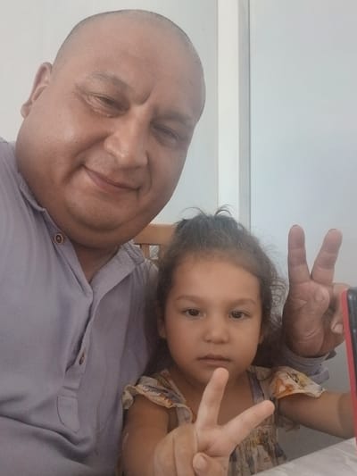 A medium shot selfie taken from a slightly high angle shows an adult male and a young girl posing together. The male, with a bald head and round face, wears a light blue collared shirt. The girl, with dark hair, is wearing a patterned dress. Both are smiling and holding up peace signs with their hands. The background appears to be a plain, light-colored wall, suggesting an indoor setting. The image was taken in Gulistan, Uzbekistan.