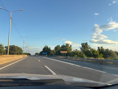 The media shows a view from a vehicle driving on a highway in Georgia, likely near Mukhrani. The road is paved and has multiple lanes, with white lines marking the lanes and a concrete barrier on the left. There are streetlights lining the highway, and trees and vegetation border the road. The sky is blue with some white, fluffy clouds, indicating it's daytime, likely late afternoon or early evening due to the warm lighting.
Two road signs are visible. One sign, further in the distance and on the left side of the road, has blue text. Another sign, closer and on the right side of the road, has brown text that reads "WINEROUTE" with an arrow pointing to the right, suggesting a scenic route related to wineries. A car is visible in the distance on the highway. The overall scene conveys a sense of travel and a journey through a scenic landscape.