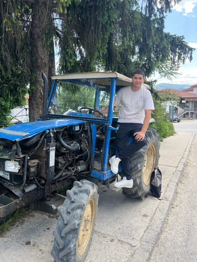 A young man is reclining on a blue tractor. The tractor is parked on the side of a road in what appears to be a rural or suburban setting. The man is wearing a white T-shirt, dark pants, and white sneakers. He is leaning against the tractor's frame. The tractor's engine is visible, and a black backpack rests on the rear tire. The background includes trees, houses, and a glimpse of a cloudy sky. The scene is in Slashten, Bulgaria.