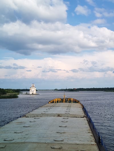 The image captures a panoramic view of the Sheksna River in Krokhino, Russia, on a partly cloudy day. In the foreground, the deck of a large barge stretches out, its surface marked by numerous circular hatches. The barge is equipped with yellow and blue railings and cargo securing mechanisms.
Dominating the mid-ground, on a small island in the river, stands the distinctive white, multi-story Church of the Nativity of the Virgin Mary. Its architecture is somewhat unconventional, resembling a lighthouse or a fortified tower with arched windows and a tiered roof. The church appears weathered, suggesting its age and exposure to the elements.
The river itself is wide and calm, reflecting the soft light of the sky. The background features a dense line of green trees along the riverbank, with a few structures visible in the distance, including what appears to be another, smaller lighthouse or navigational marker. The sky is a dramatic canvas of white and grey clouds, interspersed with patches of blue. A single bird is visible perched on the railing of the barge in the foreground.