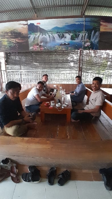 Six adult males are observed in a semi-open traditional seating area located in Keudealue Ie Puteh, Indonesia. The environment features a corrugated metal roof, wooden support structures, and wooden platforms for seating. On the floor below the seating platforms, various pairs of sandals and shoes are neatly arranged.
The men are seated on a wooden platform around a low, rectangular wooden table. All individuals are barefoot. From left to right:
1. A male in a dark long-sleeved shirt and light-colored pants sits cross-legged, holding a dark smartphone. He looks towards the camera with a neutral expression.
2. A male in a white t-shirt and blue jeans leans forward, engaged with a smartphone in his hands.
3. A male in a brown patterned shirt and dark pants smiles subtly at the camera.
4. A male in a blue collared shirt and dark pants smiles at the camera.
5. A male in a light beige polo shirt and dark jeans smiles broadly at the camera.
6. A sixth male, partially obscured by the fifth, is visible in a blue shirt, seated on the platform.
The wooden table holds multiple clear glasses containing liquid, three red-brown ceramic mugs (one with a red saucer), a red cigarette pack, a lighter, a white ceramic ashtray with cigarette butts, at least three smartphones, and a white power bank or charging device connected by a cable.
The background wall above the seating features three large rectangular prints or paintings depicting natural landscapes, with the central image prominently displaying a large waterfall. Dark mesh or netting is visible beyond the seating area, suggesting an open-air design with views to the outside greenery. The overall scene indicates a casual social gathering.