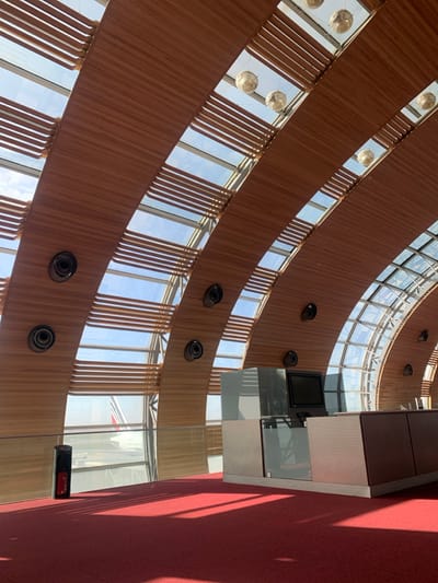 The image depicts the interior of an airport terminal in Le Mesnil-Amelot, France, likely Charles de Gaulle Airport. The scene is dominated by a distinctive arched ceiling structure, constructed from alternating bands of light-brown wooden slats and clear glass panels, through which a bright blue sky with scattered white clouds is visible. Several spherical, reflective light fixtures are suspended from the ceiling, and multiple circular dark vents or speakers are embedded into the wooden arches.
The floor is covered with a uniform vibrant red carpet, upon which strong natural light casts defined shadows from the architectural elements. In the mid-ground, a modern, angular counter or desk structure is present, featuring reflective dark and metallic panels and a dark, blank monitor screen. A low, clear glass barrier or railing runs horizontally, separating the carpeted area from large, clear windows that offer a view of an airfield.
Outside the windows, a portion of an aircraft's tail fin is discernible, displaying a white fuselage with a red and blue logo or stripe. To the left, near the glass barrier, a black cylindrical object, possibly a waste bin or fire extinguisher stand, rests on the red carpet. No individuals are present in the scene, and no discernible actions are taking place.