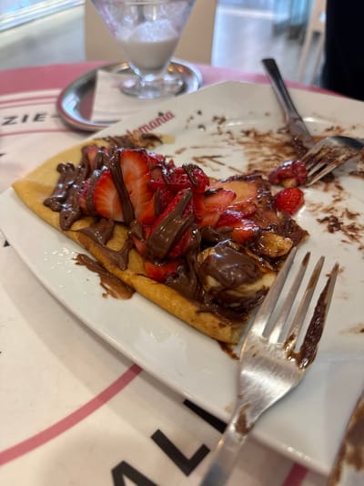 A close-up shot captures a slice of waffle topped with sliced strawberries and chocolate sauce on a white plate. The waffle appears to be folded, forming a cone-like shape. Dark chocolate sauce is generously drizzled over the strawberries and the waffle itself, with some of it spilling onto the plate. A fork rests on the plate, its tines speckled with chocolate, suggesting the dish has been partially eaten. Another fork is visible on the left side of the plate, also showing signs of use.

In the background, a glass with a clear liquid, possibly water or a light-colored beverage, is partially visible. The plate has some text and a pink stripe, with the visible words being "toria" and "ZIE". The overall impression is of a delicious dessert enjoyed at a casual dining setting. The lighting is bright, suggesting it could be daytime. The presence of cutlery and a partially consumed dessert indicates an ongoing meal or a recently finished one.