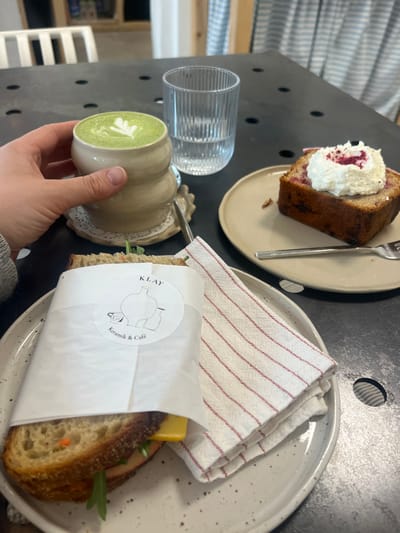 The scene depicts a cafe tabletop at KLAY Keramik & Café in Kaiserslautern, Germany. A light-skinned left hand grasps a beige, custom-shaped ceramic mug containing a green liquid with a white latte art design. This mug rests on a white, lace-patterned coaster. To its right, a clear, vertically ribbed glass holds water. On a light-colored, speckled ceramic plate, a sandwich wrapped in white paper with the "KLAY Keramik & Café" logo is visible, revealing dark bread, a slice of yellow cheese, and green and orange vegetable fillings. A folded white cloth napkin with thin red stripes lies beside the sandwich. On an adjacent, plain light-colored ceramic plate, a rectangular slice of baked goods, topped with white whipped cream and red sprinkles, is presented with a silver fork. The table surface is dark gray with circular perforations. The background is out of focus, showing blurred light-colored wooden furniture and a light blue and white striped textile.