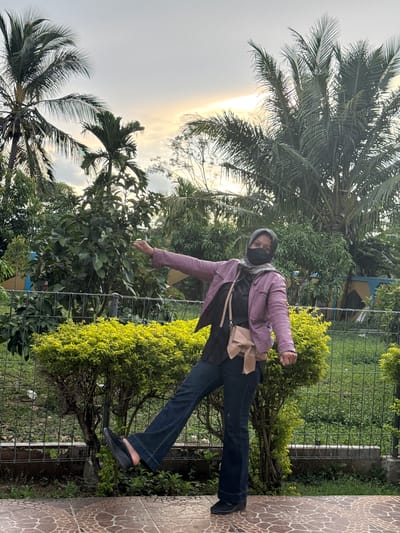 A woman stands on a tiled patio next to a fenced-off green area with lush foliage and palm trees. She is wearing a purple jacket, dark top, dark pants, a black face mask, and a grey headscarf. A light-colored crossbody bag is slung over her shoulder. The woman is looking at the camera and has her right arm extended, as if reaching for something. The sun is setting, casting a warm glow through the sky. The weather appears to be cloudy. The scene is set in Lampeuneurutblang, Indonesia.