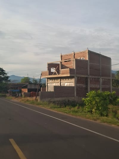 A long shot captures a three-story building under construction in Meukokuthang, Indonesia. The building, made of red brick and concrete, has an unfinished facade with exposed rebar at the top. To the left of the building are smaller structures, one with a corrugated metal roof and another with a blue tarp. A paved road with white and yellow lane markings runs diagonally across the foreground. In the background, a lush green landscape leads to distant mountains under a hazy sky.