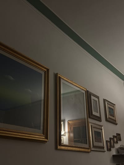 The photograph shows a wall with a collection of framed pictures hanging on it. The frames are primarily gold and various other neutral tones. The pictures vary in size. Some of the images are reflected in the glass of the frames. The wall is pale in color, with a green stripe running diagonally along the upper left edge of the frame. The scene is dimly lit. The location is Perugia, Italy.