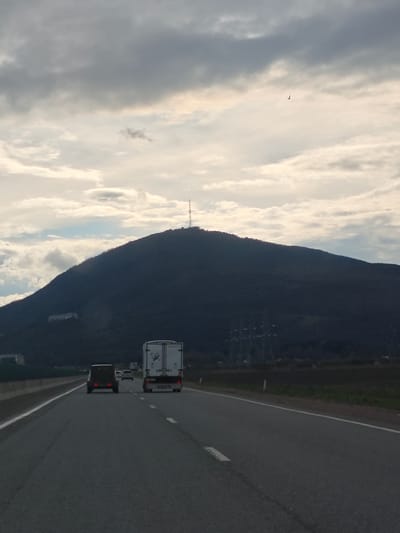 The image shows a highway with multiple vehicles traveling on it, including a semi-truck and a car. A large mountain looms in the background, with a radio tower visible at its peak. The sky is cloudy. The location is not specifically Konstantinovskaya, Russia, however the image may contain characteristics found in or around that city.